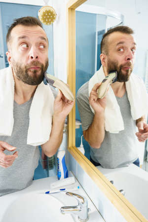 Bearded man looking at himself in mirror trimmng, shaving his beard using electric timmer razor.の写真素材