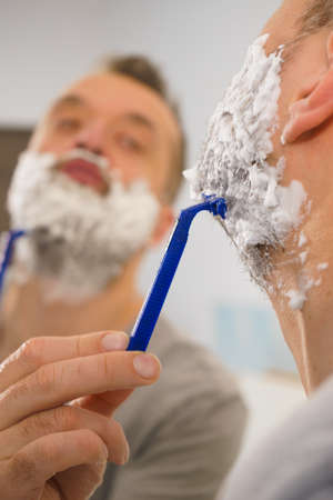 Man with lots of shaving foam on his face standing in bathroom, looking at mirror, shaving his face...