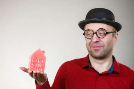 Adult man wearing funny hat and eyeglasses holding small red house model. Guy being real estate...