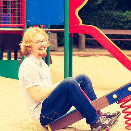 Funny nerd geek werid man sitting on swing playing and having fun on kids playground.の写真素材