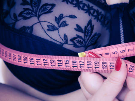 Plus size fat woman wearing black bra using tape measure to check the measurements of her big chest breasts. Closeup part of female body. Bosom, brafitting and underwear concept.の写真素材