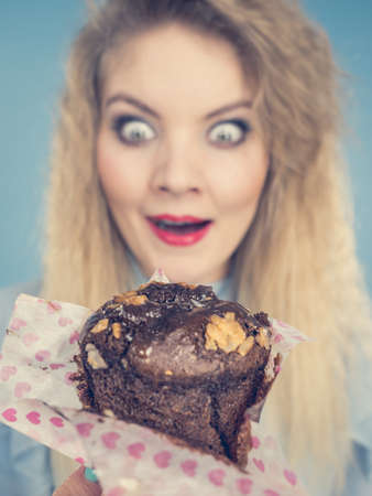 Sweet food sugar make us happy. Attractive blonde woman holds yummy chocolate cupcake in hand, open mouth, craving for cake. On blueの写真素材