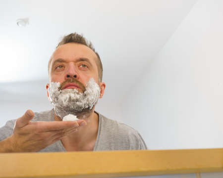 Man applying shaving foam cream on his face, standing in bathroom, looking at mirror, preparing to shave his face beard. Skincareの写真素材