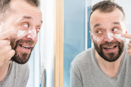 Happy funny adult guy applying moisturizer cream for male skin care. Man in front of bathroom mirror with anty aging lotion on face.の写真素材