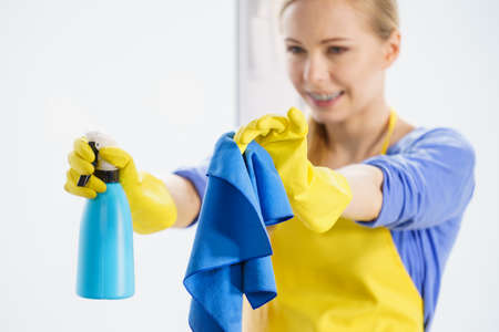 Young woman in gloves and apron washing window pane at home with rag and spray detergent. Blue...