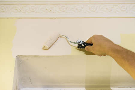 Person appplying white paint on yellow wall using roller brush. Home renovation concept.