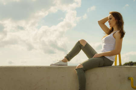Young pretty fashion model woman sitting on concrete wall wearing white tank top and olive green trousers. Female walking outdoor during warm summer weather.の写真素材