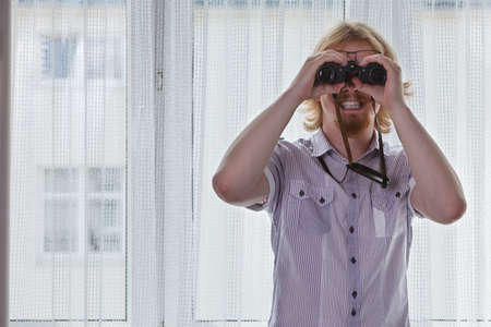 Young adult man spying at his neighbors, guy using binoculars looking through window.の写真素材