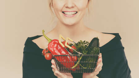 Buying good food, vegetarian products. Positive woman holding shopping basket with green red vegetables inside, recommending healthy high fibre diet, lifestyle modification, on greyの写真素材