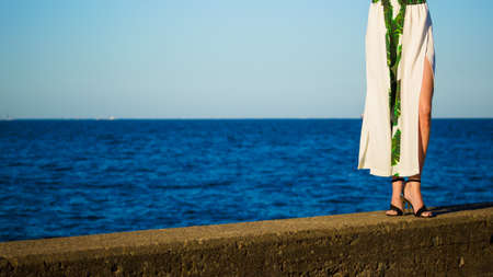 Unrecognizable woman in elegant outfit walking on seaside pier next to sea water. Female wearing...