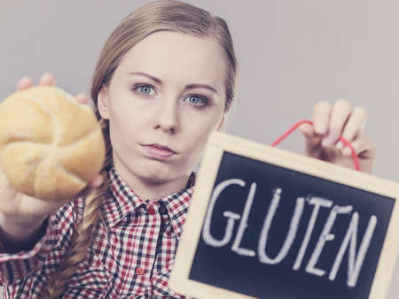 Young woman with braided hair holding small black board with gluten sign and bun bread roll. Bakery and bread allergy problem.の写真素材