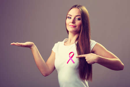Woman wih pink cancer ribbon on chest holding open hand for product text, pointing with finger. Health care, medicine and breast cancer awareness concept. Studio shot on grayの写真素材