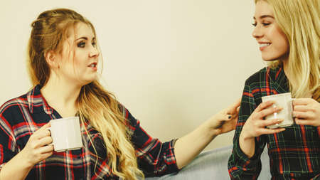 Two female friends sitting together on sofa chatting talking and drinking tea or coffee having fun...