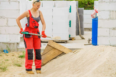 Woman working hard on construction site, using shovel digging sand soil. Partially built new home...
