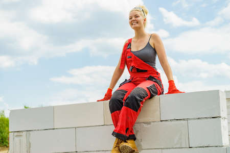 Woman wearing workwear on construction site. Female takes break from work, sitting on wall. Partially built new house early stage. Industry.の写真素材