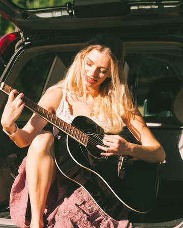 Travel vacation hitchhiking concept. Summer girl hippie style sitting on hatchback car with acoustic guitarの写真素材