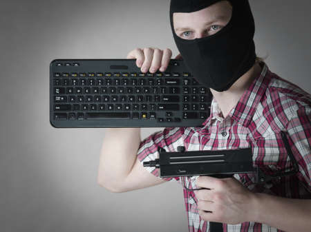 Crazy hacker man. Unrecognizable guy wearing black balaclava holding computer keyboard and gun. Hate...