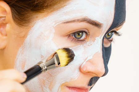 Woman with clay carbo black mask on half face applying white mud to clean skin. Girl taking care of...