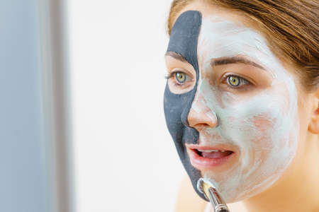 Woman with clay carbo black mask on half face applying white mud to clean skin. Girl taking care of oily complexion. Beauty procedures. Skincare.の写真素材
