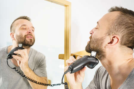 Bearded man looking at himself in mirror trimmng, shaving his beard using electric timmer razor.の写真素材