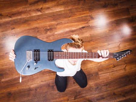 Young bearded man holding, raising his electric guitar. Hobby, music concept, on wooden floor. View from topの写真素材