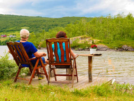 Mature couple woman and man relaxing outdoor, sitting on wooden chairs on lake fjord shore. Holidays relaxation and trip. Norway Scandinavia Europe.の写真素材