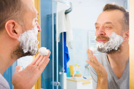 Man applying shaving foam cream on his face, standing in bathroom, looking at mirror, preparing to shave his face beard. Skincareの写真素材