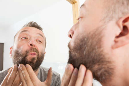 Mature adult man investigating his unshaved facial hair. Guy thinking about shaving his short beardの写真素材
