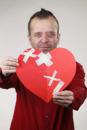 Healed love. Valentines Day concept. Adult smiling man holding big red heart fixed with plaster bandage. Male healing relationship.の写真素材