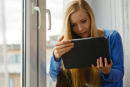 Happy young teenage woman using tablet while sitting on windowsill and relaxing surfing on internet
