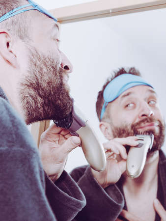 Bearded man looking at himself in mirror trimmng, shaving his beard using electric timmer razor. Guy taking care of facial hair, wearing sleepy eyemask.の写真素材
