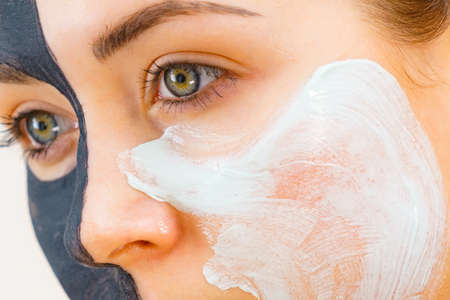 Woman with clay carbo black mask on half face applying white mud to clean skin. Girl taking care of oily complexion. Beauty procedures. Skincare.の写真素材