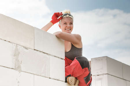 Happy blonde woman wearing dungarees about to do some work on construction site. Female sitting on wall made of airbricks taking a break.の写真素材