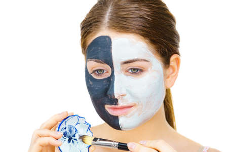 Woman with clay carbo black mask on half face applying white mud to clean skin. Girl taking care of oily complexion. Beauty procedures. Skincare.の写真素材