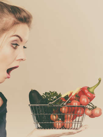 Adult woman do not like to eat raw food, questioning healthy lifestyle recommendations, origin vegetagles. Female holding small shopping basket with products, displeased shocked face expressionの写真素材