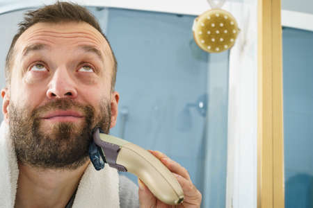 Bearded man looking at himself in mirror trimmng, shaving his beard using electric timmer razor.の写真素材