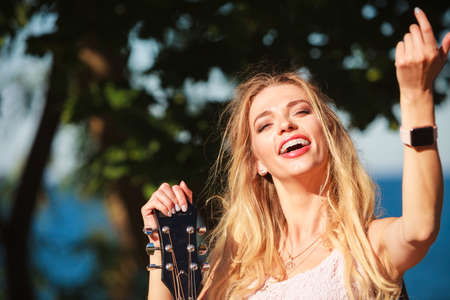 Blonde woman having acoustic guitar. Female playing music on seaside