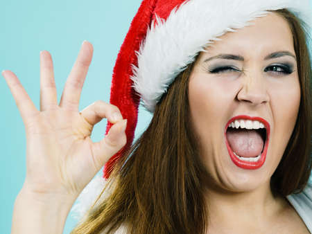 Happy joyful girl wearing santa hat giving thumb up ok sign, on blue. Christmes time.の写真素材