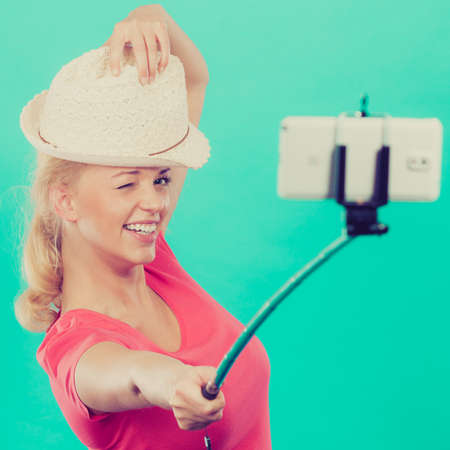 Technology, modern photography, confidence conept. Happy attractive adult blonde woman with sun hat taking funny picture of herself with smartphone on selfie stick.の写真素材