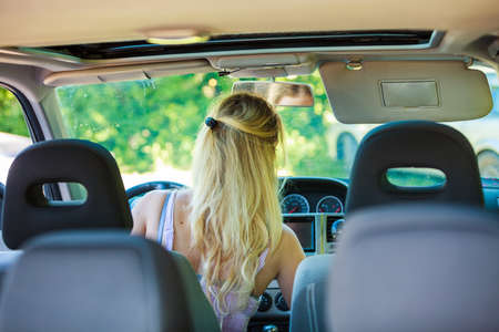 Young blonde woman driving bad car, doing something in auto, view from rear seatの写真素材