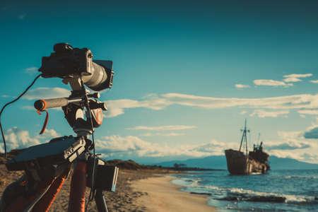 Professional camera on tripod taking picture film video from coastline with rusty shipwreckの写真素材