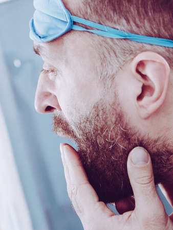 Funny adult man wearing eyemask on forhead having troubles with waking up. Standing in front of mirror looking at his face, yawning, being tired and sleepy.の写真素材