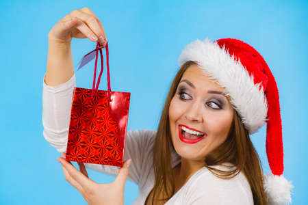 Young adult woman is happy to give Christmas gifts. Female wearing santa claus hat holding present red gift bag, on blueの写真素材