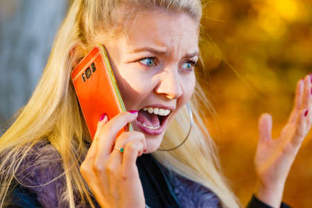 Blonde woman walking in autumn park, using mobile phone. Angry female talking on smartphone.の写真素材
