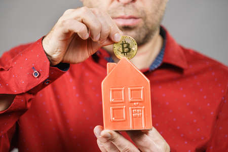 Man paying his house rent using cryptocurrency. Guy holding model of red home and bitcoin coin.の写真素材