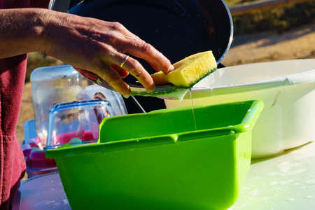 Mature woman washing up dishes in bowl on fresh air. Dishwashing outdoor on camping site, sea shoreの写真素材
