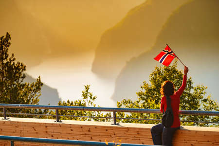 Tourist woman with norwegian flag enjoying fjord Aurlandsfjord view from Stegastein viewing point. National tourist route Aurlandsfjellet, Norwayの写真素材