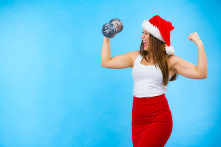 New Year fitness and building muscles resolution. Woman in santa claus hat lifting dumbbells weights, on blue. Goal achievement in training.の写真素材