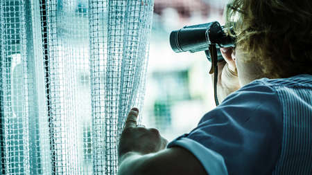 Young adult man spying at his neighbors, guy using binoculars looking through window.