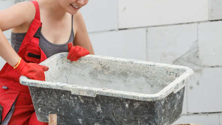 Woman wearing dungarees working on construction site of her home. Mixing cement in bowl preparing...